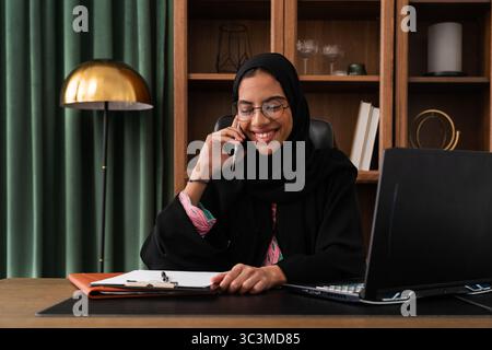 Geschäftsfrau aus dem Nahen Osten, die Abaya trägt und im Büro arbeitet - Geschäftsfrau aus emirat in traditioneller Abaya und Hijab lächelt, während sie auf dem p Stockfoto