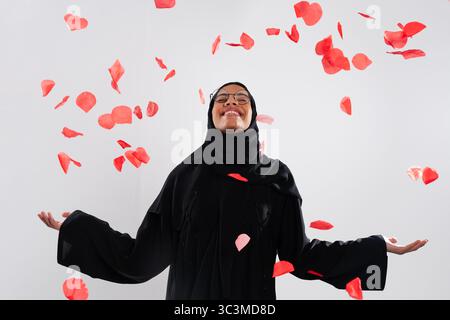 Arabische Frau, die eine traditionelle Abaya und Hijab trägt, lächelt mit offenen Armen, während rote Rosenblätter in einem Studio um sie fallen und Schönheit feiern. Stockfoto