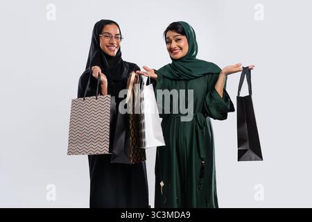 Zwei glückliche Frauen aus emirat, die traditionelle Abayas tragen, zeigen ihre Einkaufstaschen in einem Studio und geben sich als Freunde aus, die ihre Einkäufe genießen Stockfoto