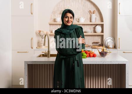 Eine Emiratin in traditioneller Abaya steht in ihrer modernen Küche, lächelt mit überkreuzten Armen, neben frischem Gemüse und einer Teekanne, die Cu verkörpert Stockfoto