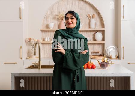 Die Emiratin, die eine traditionelle Abaya trägt, steht mit überkreuzten Armen in ihrer modernen Küche und lächelt selbstbewusst, während sie sich auf die Zubereitung einer Mahlzeit vorbereitet Stockfoto