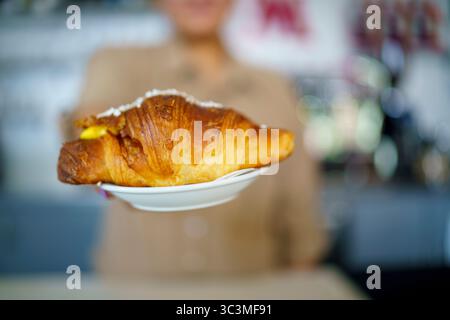 Köstliches, frisch gebackenes Golden Croissant gefüllt mit cremigem Pudding, präsentiert auf einem weißen Teller von einer lächelnden Person in einer gemütlichen Café-Umgebung Stockfoto