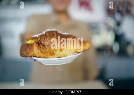 Köstliches goldenes Croissant gefüllt mit cremigem Pudding, serviert auf einem weißen Teller von einer verschwommenen Person Stockfoto