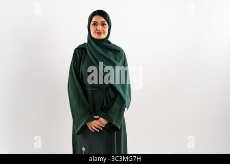 Eine selbstbewusste Frau aus dem Nahen Osten, die eine traditionelle grüne Abaya und einen Hijab trägt, posiert anmutig in einem Studio und zeigt ihre Eleganz und Kultur Stockfoto