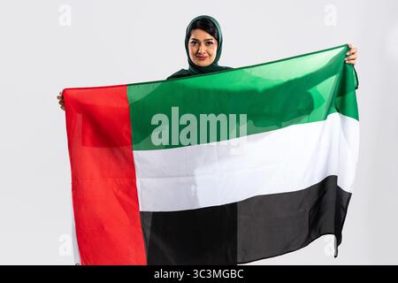 Die Emiratin trägt einen Hijab und hält stolz die Nationalflagge der vereinigten arabischen emirate in einem Studio-Setting, in dem sie ihren Patriotismus und ihre Nationalflagge zeigt Stockfoto