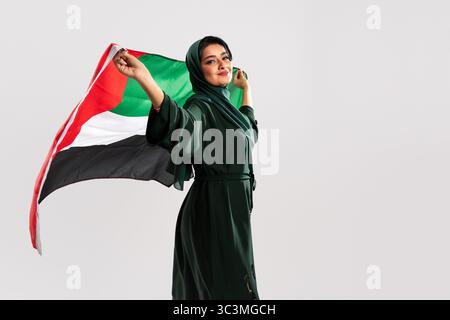 Die Emiratin trägt eine grüne Abaya und einen Hijab und hält stolz die Flagge der vae in einem Studio-Setting und verkörpert nationalen Stolz und kulturelle Identität Stockfoto