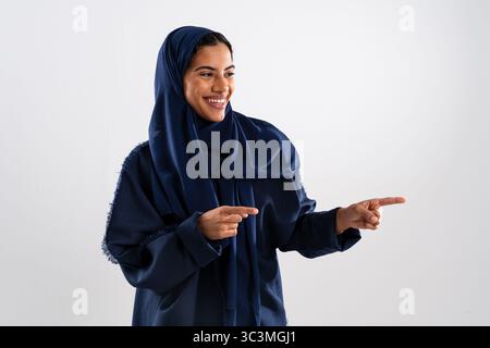 Studio-Porträt einer fröhlichen jungen muslimischen Frau, die einen blauen Hijab und Abaya trägt und mit ihren Zeigefingern etwas anzeigt, während sie lächelt und schaut Stockfoto