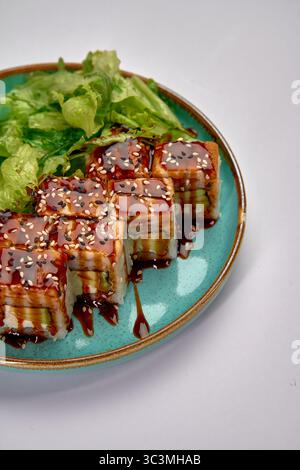 Ein köstliches Arrangement von Sushi-Brötchen mit süßer Sauce und Sesamsamen, serviert zusammen mit frischem, knusprigem Salat auf einem türkisfarbenen Teller. Stockfoto