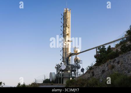 Ein hoher Telekommunikationsturm mit verschiedenen Antennen und Schüsseln steht auf einem gesicherten Hügel vor einem klaren blauen Himmel. Stockfoto