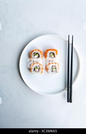 Blick von oben auf ein kalifornisches Brötchen mit frischem Lachs, serviert auf einer weißen Keramikplatte neben dunklen Essstäbchen auf einer strukturierten, hellweißen Oberfläche. Stockfoto