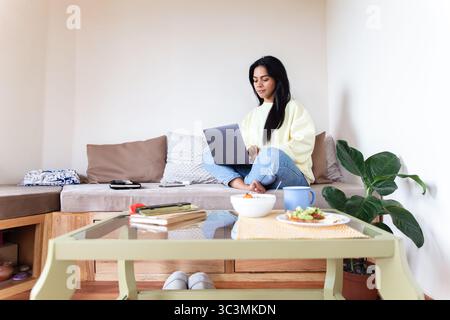 Eine Lateinfrau sitzt zu Hause bequem auf ihrem Sofa, arbeitet an ihrem Laptop und genießt einen ruhigen Tag. Die Szene fängt den modernen Alltag und das Leben ein Stockfoto
