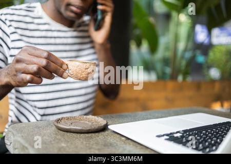 Ein Mann sitzt in einem gemütlichen Café und hält eine Kaffeetasse, während er ein Telefongespräch führt. Ein Laptop steht offen auf dem Tisch, was eine Mischung aus Arbeit und Familie andeutet Stockfoto