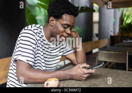 Ein Mann sitzt an einem Café-Tisch, in sein Smartphone eingesogen. Er ist lässig in einem gestreiften Hemd gekleidet und genießt seine Kaffeepause in entspannter und einladender Atmosphäre Stockfoto