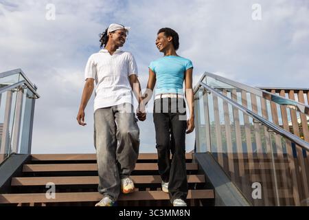 Ein fröhliches, queeres Paar geht an einem sonnigen Sommertag Hand in Hand eine Treppe hinunter, umgeben von klarem Himmel. Sie lächeln und erscheinen in einem Moment Stockfoto