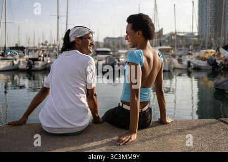 Ein queeres Paar genießt einen sonnigen Sommertag auf einem Yachthafen, umgeben von Booten. Sie teilen einen fröhlichen Moment, lächeln und entspannt, mit einem malerischen Wasser Stockfoto