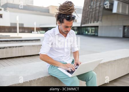 Ein Italiener mit Dreadlocks sitzt draußen auf einer Betonbank und konzentriert sich auf seinen Laptop. Die Umgebung ist ein modernes Stadtgebiet in Spanien, das trave reflektiert Stockfoto