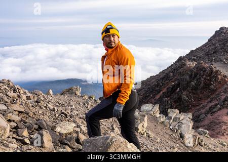 Ein Alleinwanderer in lebendiger Ausrüstung steht selbstbewusst auf einem felsigen Bergpfad und blickt über ein Wolkenmeer und symbolisiert Entdeckungsreise, Freiheit und Stockfoto
