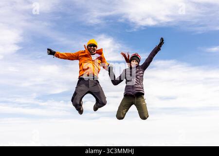 Ein Mann und eine Frau springen energisch in die Luft, umgeben von einer atemberaubenden Bergkulisse. Ihr abenteuerlicher Geist und ihre Freude sind offensichtlich, wenn sie sich einfühlen Stockfoto