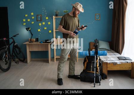 Junge Mann, der die Tour online mit dem Handy Bucht, während er die Wanderung vorbereitet und dabei Reisepass und Flugtickets zu Hause hält. Konzept von Trekking, Wandern und Stockfoto