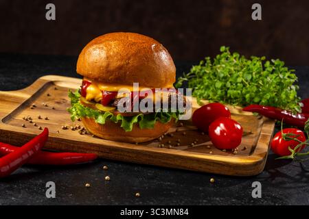 Köstlicher Cheeseburger auf einem Holzbrett mit Salat, Cheddar, Ketchup und saftigem Rindfleisch, umgeben von frischen Kirschtomaten und Stockfoto
