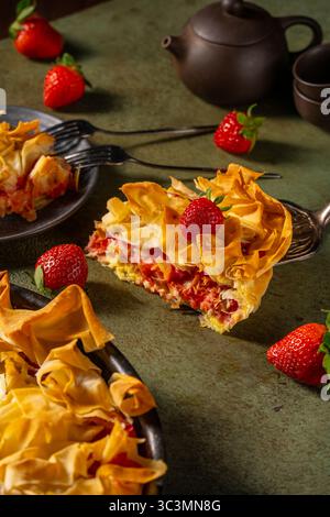 Köstlicher Erdbeerkuchen mit knusprigem Phyllo-Teig wird kunstvoll präsentiert, zusammen mit frischen Erdbeeren und eleganten Teesorten auf einem rustikalen Tisch. Stockfoto