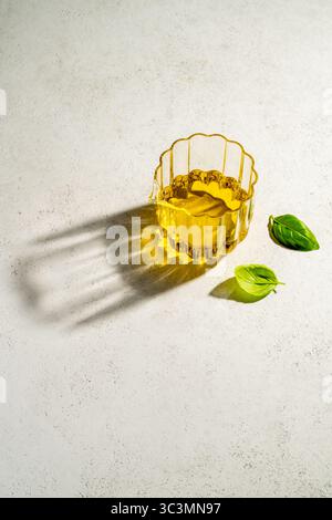 Von oben liegt ein mit goldenem Olivenöl gefülltes Glas auf einem strukturierten weißen Hintergrund neben frischen, leuchtend grünen Basilikumblättern, die einen weichen Schatten bilden Stockfoto
