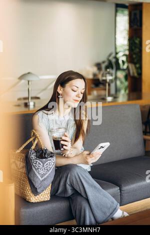 Eine Frau sitzt bequem auf einem Sofa in einem gemütlichen Café, hält einen Eiskaffee in der einen Hand und ein Smartphone in der anderen und steht für Entspannung und Moderne Stockfoto