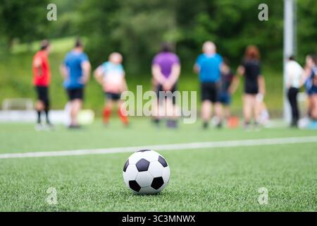 Nicht erkennbare Personen, die auf einem grünen Fußballfeld stehen, mit einem schwarz-weißen Ball im Vordergrund. Die Umgebung ist an einem sonnigen Tag mit üppiger Vegetation im Freien Stockfoto