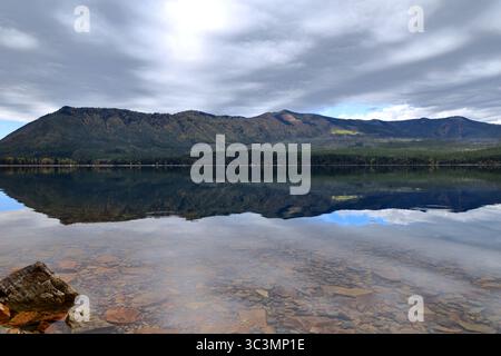 Das mit Kieselsteinen übersäte Seebad glitzert in kristallklarem Wasser und reflektiert sonnenbeschienene Hügel und Wolken über dem Lake McDonald’s ruhige, unberührte Weite. Stockfoto