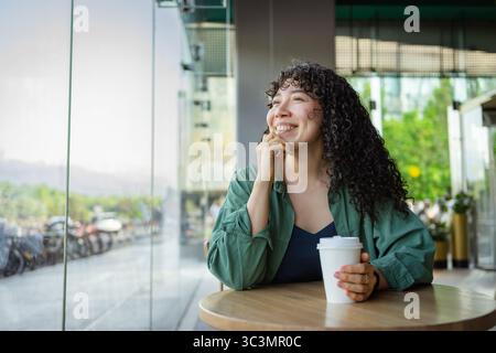 Eine junge Frau mit lockigen Haaren sitzt an einem Café-Tisch und lächelt zufrieden, während sie eine Kaffeetasse hält. Sonnenlicht strömt durch das große Fenster und erzeugt einen Stockfoto