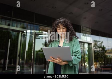 Eine junge Frau mit lockigen Haaren steht vor einem modernen Gebäude und benutzt aufmerksam ihren Laptop. Sie trägt ein grünes Hemd und konzentriert sich auf die Arbeit Stockfoto