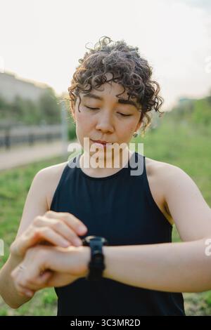 Lockige junge Frau in sportlicher Kleidung überprüft ihre Smartwatch, während sie sich im Freien aufhält. Sie wirkt fokussiert und illustriert moderne Technolo Stockfoto