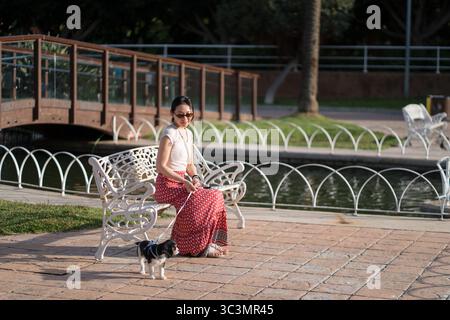 Eine Asiatin genießt den Park mit ihrem Kavalier King Charles Spaniel Welpen. Die Sonne scheint hell und unterstreicht ihre freudige Begleitung Stockfoto