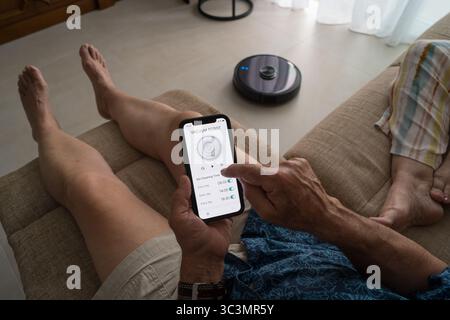 Ein älterer Mann steuert mühelos einen intelligenten Roboter-Vakuum vom Sofa aus mit einer Smartphone-App. Die intuitive Oberfläche ermöglicht eine einfache sche Stockfoto