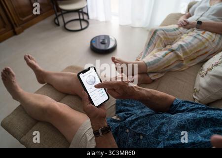 Ein älterer Mann steuert mühelos einen intelligenten Roboter-Vakuum vom Sofa aus mit einer Smartphone-App. Die intuitive Oberfläche ermöglicht eine einfache sche Stockfoto