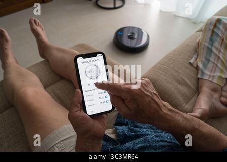 Ein älterer Mann steuert mühelos einen intelligenten Roboter-Vakuum vom Sofa aus mit einer Smartphone-App. Die intuitive Oberfläche ermöglicht eine einfache sche Stockfoto