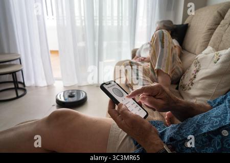 Ein älterer Mann steuert mühelos einen intelligenten Roboter-Vakuum vom Sofa aus mit einer Smartphone-App. Die intuitive Oberfläche ermöglicht eine einfache sche Stockfoto