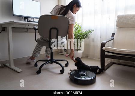 Eine asiatische Frau arbeitet in ihrem Heimbüro, während ein kleiner Hund in der Nähe liegt. Ein Roboter-Vakuum bewegt sich über den Boden und demonstriert so eine moderne und effiziente Funktion Stockfoto