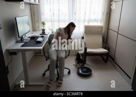 Eine asiatische Frau interagiert mit ihrem Hund in einem modernen Heimbüro, begleitet von einem Roboter-Vakuum. Die gemütliche Einrichtung umfasst einen Schreibtisch, einen Computer und eine Komfortanlage Stockfoto