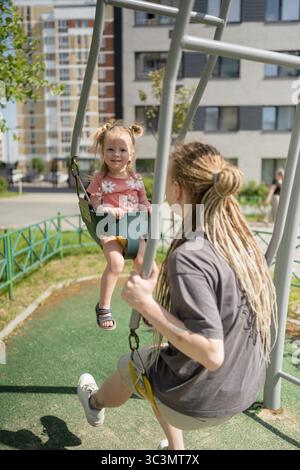 Ein freudiger Moment, als Mutter und Tochter auf einer Schaukel in einem lebhaften Park spielen. Das Kind lächelt hell und genießt den sonnigen Tag, während es sanft p Stockfoto