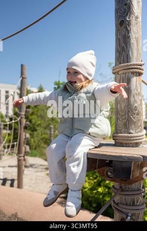 Ein glückliches Kind genießt einen Tag im Freien und sitzt spielerisch auf einem hölzernen Spielplatz. In einem gemütlichen Winteroutfit tragen sie den sonnigen Tag mit Stockfoto