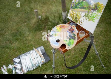 Eine Künstlerpalette und Leinwand sind auf einer Staffelei in einem grünen Park aufgestellt. Malerröhren und Pinsel liegen in der Nähe und sind bereit für eine farbenfrohe Landschaftsszene Stockfoto