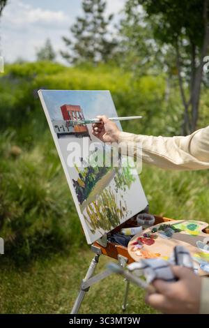 Ein Künstler malt mit einem Pinsel eine farbenfrohe Landschaft auf eine Leinwand, vor einer üppigen Kulisse im Freien. Die Szene fängt das Wesen der Kreativität und ein Stockfoto
