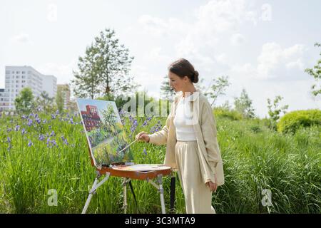 Eine Frau malt eine lebendige Landschaftsszene auf einer Staffelei, umgeben von üppigem Grün und lila Wildblumen, und fängt die natürliche Schönheit an einem sonnigen Tag i ein Stockfoto