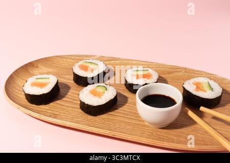 Sushi-Brötchen mit Lachs, Gurke und Reis auf einer Holzplatte. Eine kleine Tasse Sojasauce und Essstäbchen sind auf einem sanften rosa Hintergrund angeordnet. Stockfoto