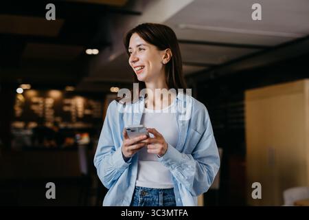 Eine Frau in einem lässigen blauen Hemd und weißem Oberteil steht lächelnd und hält ein Smartphone. Sie arbeitet aus der Ferne in einem gemütlichen Café Stockfoto