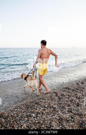 Ein blinder Mann spaziert mit seinem Begleithund an einem Sandstrand entlang und genießt die ruhige Umgebung am Meer. Betonung der Verbundenheit und Freiheit einer integrativen li Stockfoto