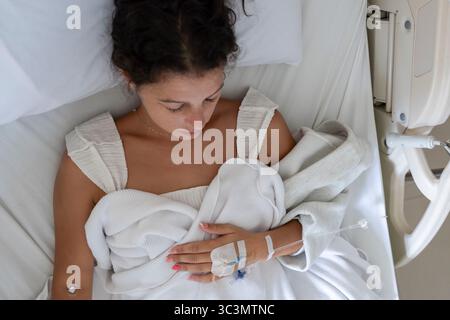 Eine kranke junge Frau liegt in einem Krankenhausbett in Bali, eingewickelt in eine weiße Decke. Eine IV ist an ihrer Hand angebracht, was eine medizinische Behandlung für sie andeutet Stockfoto