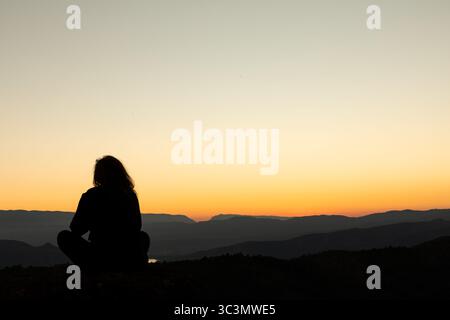 Rückansicht einer nicht erkennbaren Frau, die beim Meditieren sitzt und den ruhigen Herbstuntergang über den majestätischen Pyrenäen genießt. Der Horizont ist wunderschön Stockfoto