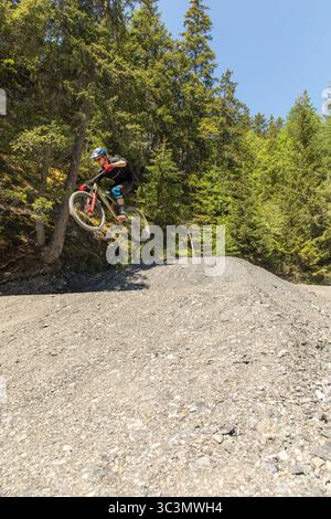 Von unten steigt ein männlicher Mountainbiker schnell einen Schotterweg in den Alpen ab, umgeben von üppigem Grün. Ausgestattet mit Sicherheitsausrüstung, demonstriert er Stockfoto